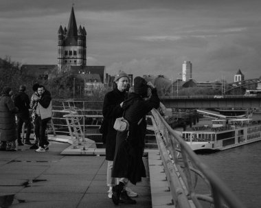 Cologne, Germany - October 27, 2023: Couples and small groups lean on the bridge railing, gazing across the calm river. The historic skyline in the background blends beautifully with the industrial bridges, underlining Colognes dynamic charm. clipart