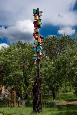 Fotoğraf üst üste yığılmış birçok renkli kuş evlerinin olduğu uzun bir tahta direği gösteriyor. Direği çevreleyen yeşil ağaçlar, çimenler ve arka planda çatısı olan bir kuyu vardır..