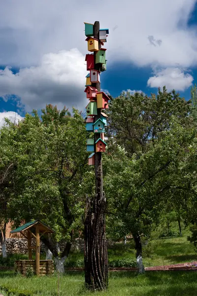 Fotoğraf üst üste yığılmış birçok renkli kuş evlerinin olduğu uzun bir tahta direği gösteriyor. Direği çevreleyen yeşil ağaçlar, çimenler ve arka planda çatısı olan bir kuyu vardır..
