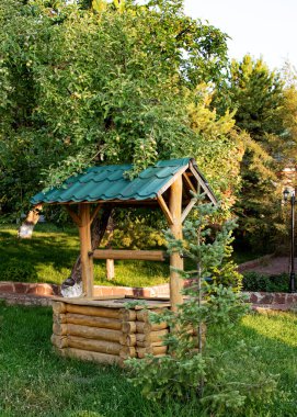 Fotoğrafta, bahçesinde yeşil çatılı dekoratif ahşap bir kuyu görülüyor. Etrafı ağaçlar, çimenler ve sıcak akşam ışığında yıkanmış genç bir ladin fidanı ile çevrili..