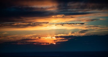 Fotoğraf, parlak turuncu ve altın güneş ışınlarının katmanlı bulutları aşarak gün batımını yakalar. Ufuk karanlık ve gökyüzü çarpıcı ışık ve zengin renklerle dolu..