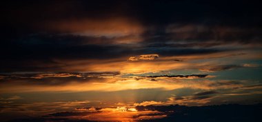 Fotoğraf, parlak turuncu ve altın güneş ışınlarının katmanlı bulutları aşarak gün batımını yakalar. Ufuk karanlık ve gökyüzü çarpıcı ışık ve zengin renklerle dolu..