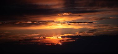 Fotoğraf, parlak turuncu ve altın güneş ışınlarının katmanlı bulutları aşarak gün batımını yakalar. Ufuk karanlık ve gökyüzü çarpıcı ışık ve zengin renklerle dolu..