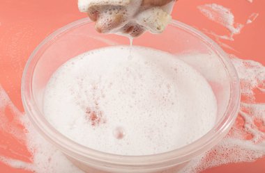 A Basin with Soapy Water, Hand Holds Soapy Sponge with Foam on Pink Background, Copy Space