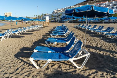 Mavi şemsiyeli plaj yatakları, Tenerife sahil beldesinde kumlu plajlarda sandalyeler, sahil kenti dinlenme alanı, dinlenme, sabah ışığında boş sahil sandalyeleri, okyanus manzarası.