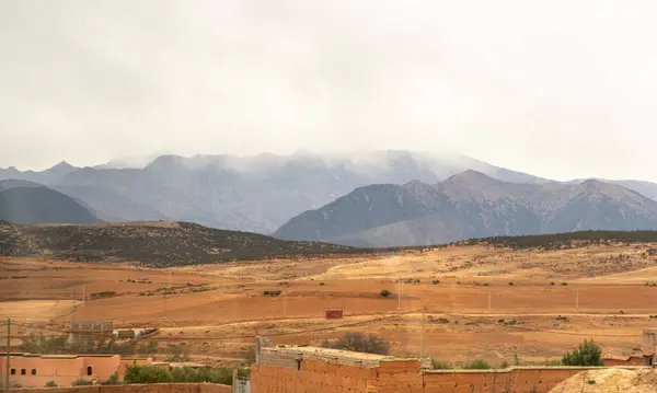 Stock image Arid Moroccan landscape near Marrakech, sparse vegetation, rolling hills, dry terrain, distant mountains, scattered trees, remote buildings, winding road, desert-like, rural scenery