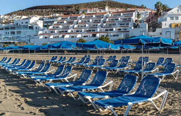 stock image Beach sunbeds with blue umbrellas, chairs on sandy beach on Tenerife seaside resort, coastal town leisure area, relaxation, empty coastal chairs in morning light, oceanfront