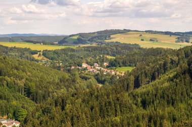 Tepeler, ormanlar ve parçalı bulutlu gökyüzünün altındaki tarlalarla çevrili yeşil bir vadide yuva yapmış küçük bir köy manzarası.