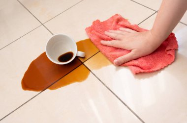 Dökülmüş kahve, kadın fayanslı zemine kahve lekesini siliyor, beyaz bardakla karışık sızıntı, kahverengi içki dökülmüş temizlik.