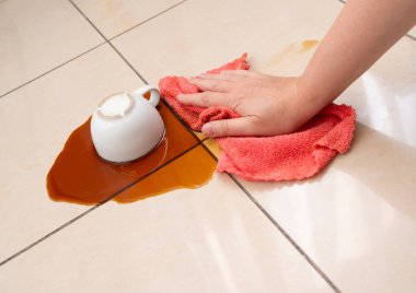 Dökülmüş kahve, kadın fayanslı zemine kahve lekesini siliyor, beyaz bardakla karışık sızıntı, kahverengi içki dökülmüş temizlik.