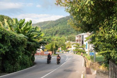 Beau Vallon, Seyşeller, 31 Ocak 2025: Beau Vallon, Seyşeller 'de iki motorcu, tropikal yeşillik ve dağ manzarasıyla çevrili virajlı bir yolda ilerliyor
