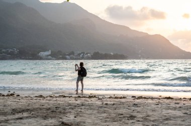 Beau Vallon, Seyşeller, 2 Şubat 2025: Sırt çantalı bir turist, tropik bir sahil editörünün kumlu sahilinde çıplak ayakla gün batımını yakalar.