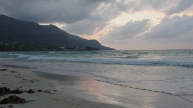 Seyşeller 'deki Beau Vallon plajında gün batımı ve kıyı manzarası, dramatik gökyüzü altında dalgalar, tepelerin siluetleri ve tropikal kıyı şeritleri yer alıyor.