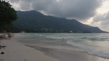 Seyşeller 'deki Beau Vallon plajında gün batımı ve kıyı manzarası, dramatik gökyüzü altında dalgalar, tepelerin siluetleri ve tropikal kıyı şeritleri yer alıyor.