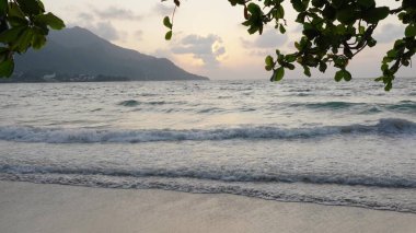 Seyşeller 'deki Beau Vallon plajında gün batımı ve kıyı manzarası, dramatik gökyüzü altında dalgalar, tepelerin siluetleri ve tropikal kıyı şeritleri yer alıyor.