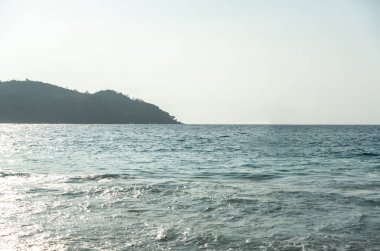 Sakin Hint Okyanusu 'nun geniş manzarası. Güneş ışığı, Berrak bir tropikal gökyüzünün altındaki yemyeşil Seyşeller adası kıyısında hafif dalgalardan yansıyor.