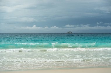 Tropik turkuaz okyanus dalgaları Seyşeller 'deki kumlu sahilde kırılıyor bulutlu gökyüzünün altında uzak bir ada doğal güzelliği, yaz cennetini ve huzurlu kıyı manzarasını yakalıyor.