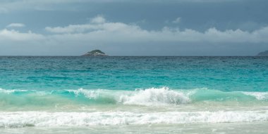 Tropik turkuaz okyanus dalgaları Seyşeller 'deki kumlu sahilde kırılıyor bulutlu gökyüzünün altında uzak bir ada doğal güzelliği, yaz cennetini ve huzurlu kıyı manzarasını yakalıyor.