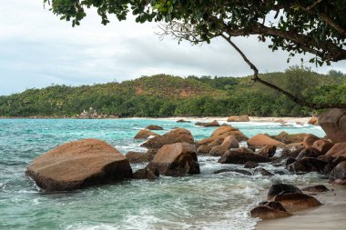 Seyşeller adasındaki tropik bir sahil boyunca granit kayalar ve turkuaz su, arka planda yemyeşil tepeler, seyahat ve doğa temaları için ideal doğal cennet manzarası.