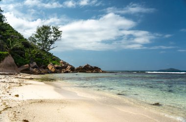 La Digue Adası 'ndaki beyaz plaj deniz çim yığınları, temiz sığ okyanus suyu, küçük dalgalar ve sıcak bir tropikal günde güçlü güneş ışığı ile kaplıdır.