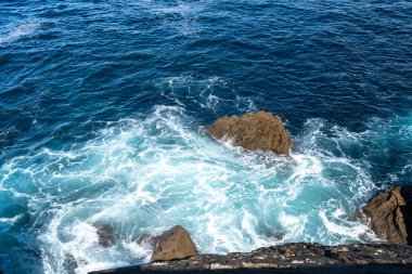 Dalgalar kıyı kayalarına çarpar ve görünür hareket ve yüzey dokusuyla berrak mavi okyanus suyuna köpük ve sprey püskürtür.