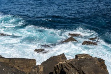 Dalgalar aşınmış delikler ve gelgit havuzlarıyla kayalık bir sahile çarpar ve deniz kenarında gezginler için vahşi ve manzaralı bir manzara yaratır.