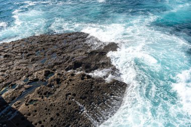Okyanus su sıçramaları ve köpüklerle parıldıyor. Dalgalar kayalara çarpar ve suda oynayan dev gibi ses çıkarırlar.