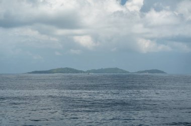 Seyşeller, Praslin ve La Digue arasındaki uzak adaların deniz yolculuğu sırasında bir feribottan yakalanan bulutlu gökyüzünün altında bir yelkenliyle okyanusta görüntüsü.