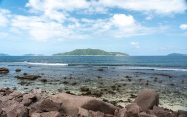 La Digue ve Praslin, Seyşeller arasındaki bulutlu gökyüzünün altında ufukta görünen granit kıyı kayaları ve okyanus dalgaları kıyı ve deniz manzaralarını gösteriyor.