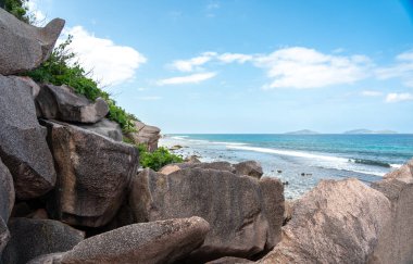 La Digue Adası, Seyşeller 'in kayalık granit sahil şeridinden, Praslin' in uzak adaları ile okyanusa bakan, parçalı bulutlu bir gökyüzü altında, doğal kıyı manzarasını gösteren
