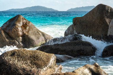 Okyanus ve deniz dalgaları mavi gökyüzünün altında sahil kıyısı boyunca kaya ve suya çarpar. Yaz mevsiminde kayalık kayalıklar ve uzak adalarla tropikal kıyılarda su ve köpük yükselir.