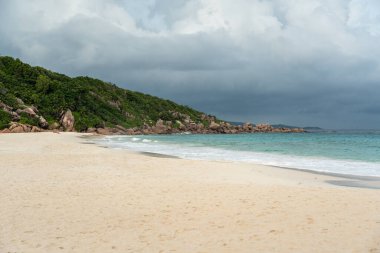 Seyşeller 'deki La Digue adasındaki beyaz bir kumsal üzerinde kara fırtına bulutları toplanıyor. Mavi okyanus dalgaları kıyıya ulaşır tropikal kayaların ve yeşilliklerin yanında