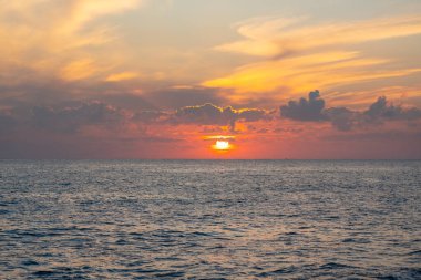 Akdeniz üzerinde gün batımı yumuşak dalgalar ve parlayan bulutlarla. Sıcak renkler suyu yansıtır ve doğal bir eğilim yaratır.
