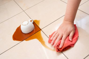 Dökülmüş kahve, kadın fayanslı zemine kahve lekesini siliyor, beyaz bardakla karışık sızıntı, kahverengi içki dökülmüş temizlik.