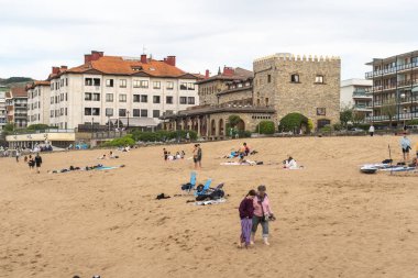 Zarautz, İspanya, 4 Nisan 2024: İnsanlar tarihi bir taş bina ve kıyı otelleri yakınlarındaki Zarautz, İspanya 'nın kumlu sahillerinde dinleniyor. Kültür ve eğlence karışımı, editör imajı.