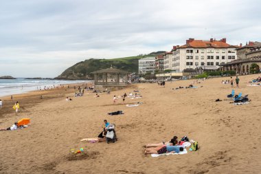 Zarautz, İspanya, 4 Nisan 2024: Aileler İspanya 'nın Zarautz plajında tarihi bir pavyon ve kıyı binalarının yakınında güneşleniyorlar. Huzurlu bir deniz kenarı inzivası, editör imajı