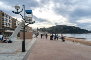 Zarautz, İspanya, 4 Nisan 2024: İnsanlar İspanya 'da sahil ve orman tepeleri yakınlarındaki Zarautz gezinti alanı boyunca yürüyorlar. Okyanus ve günbatımı manzaralı sakin bir sahil manzarası, editoryal görüntü.