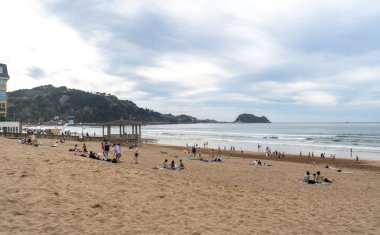 Zarautz, İspanya, 4 Nisan 2024: İnsanlar okyanus dalgaları, bir çardak ve bulutlu bir gökyüzü altındaki uzak tepelerin, editör görüntüsünün olduğu Zarautz kumsalının tadını çıkarıyorlar