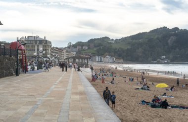 Zarautz, İspanya, 4 Nisan 2024: İnsanlar Zarautz 'da deniz kenarında gezinirken, diğerleri tepeler ve şehir binaları yakınlarındaki kumsalda dinleniyor.