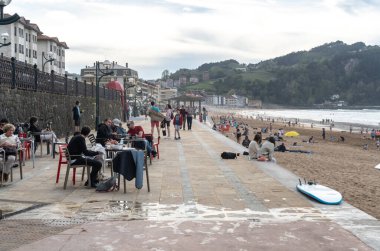 Zarautz, İspanya, 4 Nisan 2024: İnsanlar deniz kenarındaki kafelerde dinleniyor ve Zarautz 'daki gezinti alanında yürürken, diğerleri yeşil tepelerin yakınındaki sahilin tadını çıkarıyor, editoryal görüntü