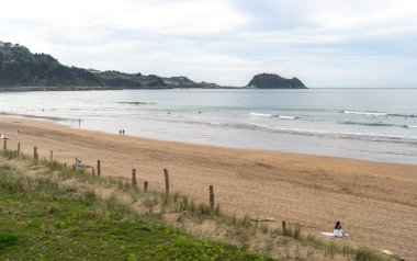 Zarautz, İspanya, 4 Nisan 2024: İnsanlar Zarautz 'da kumlu sahil boyunca yürürken sörfçüler kayalık adaların yakınında dalgalar yakalıyor.