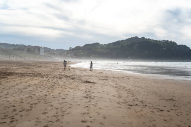 Zarautz, İspanya, 4 Nisan 2024: Çocuklar puslu Zarautz plajında top oynarken dalgalar geliyor ve insanların siluetleri tepelerin yakınındaki sahil şeridinde yürüyor, editoryal görüntü