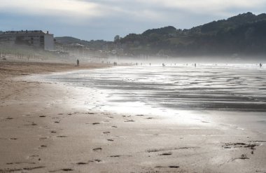 Zarautz, İspanya, 4 Nisan 2024: Akşam sisi Zarautz plajını kaplarken, insanların siluetleri Bask tepeleri yakınlarındaki yumuşak ışık altında yürüyor.