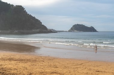Zarautz, İspanya, 4 Nisan 2024: Akşamları yumuşak dalgalar ve kıyıda yürüyen bir aile ile birlikte geniş bir Zarautz plajı, tepeler ve bir ada, editoryal görüntü