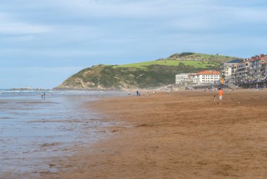 Zarautz, İspanya, 4 Nisan 2024: Zarautz plajı yeşil tepeler ve Bask Ülkesi 'ndeki kıyı binaları yakınlarındaki ıslak kum üzerinde yürüyen insanlarla geniş bir alana yayılır.