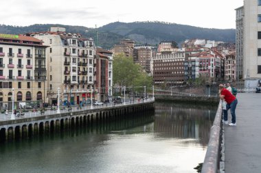 Bilbao, İspanya, 4 Nisan 2024: İnsanlar Bilbao 'da nehir kenarındaki korkulukların yanında duruyor ve arka planda şehir binalarının ve uzak tepelerin yansımaları var.
