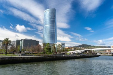 Bilbao, Fransa, 28 Mart 2024: Iberdrola Kulesi ve Bilbao 'da yaya köprüsü, palmiye ağaçları ve kent parkı ile modern nehir kenarı gezinti alanı