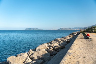 Palermo, İtalya, 18 Kasım 2024: Palermo 'nun deniz, dağlar ve Monte Pellegrino ile kayalık gezinti alanında oturan kişi, sakin Akdeniz Körfezi, editoryal görüntü