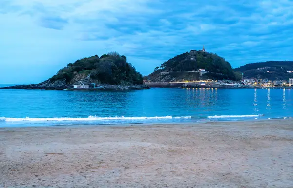 San Sebastian, İspanya, 17 Nisan 2024: San Sebastian körfezindeki Urgull ve Santa Clara adalarının sakin deniz, kumlu plaj ve şehir ışıkları manzaralı akşam manzarası