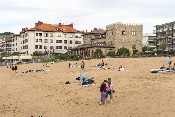Zarautz, İspanya, 4 Nisan 2024: İnsanlar tarihi bir taş bina ve kıyı otelleri yakınlarındaki Zarautz, İspanya 'nın kumlu sahillerinde dinleniyor. Kültür ve eğlence karışımı, editör imajı.